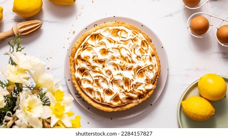 French lemon tart with churned meringue on marble table with some lemons, eggs, and spring yellow flowers in the corner, top view, horizontal image - Powered by Shutterstock