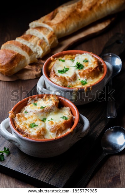French onion soup in soup crocks with\
a bread baguette in behind, against a dark\
background.
