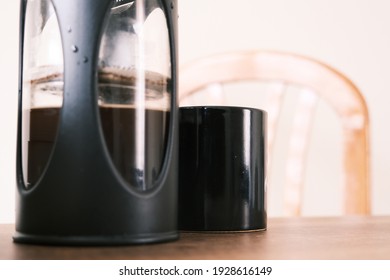 French Press Coffee Maker And It Is Half Filled With Arabic And Black Caffe Together With Black Caffe Pot With Upper Side Of Wooden Chair. All Things Standing On Wooden Table.