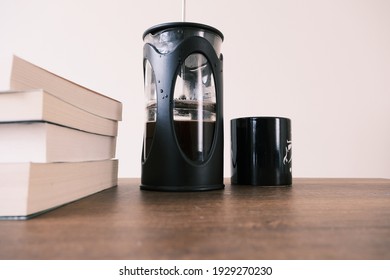 French Press Coffee Maker And It Is Half Filled With Arabic And Black Caffe Together And Also Bundle Of Books With Black Caffe Pot With Upper Side Of Wooden Chair. All Things Standing On Wooden Table.