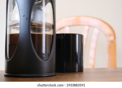 French Press Coffee Maker And It Is Half Filled With Arabic And Black Caffe Together With Black Caffe Pot With Upper Side Of Wooden Chair. All Things Standing On Wooden Table.