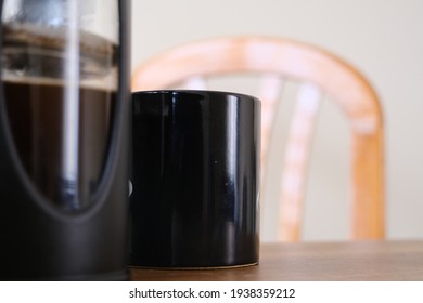 French Press Coffee Maker And It Is Half Filled With Arabic And Black Caffe Together With Black Caffe Pot With Upper Side Of Wooden Chair. All Things Standing On Wooden Table.