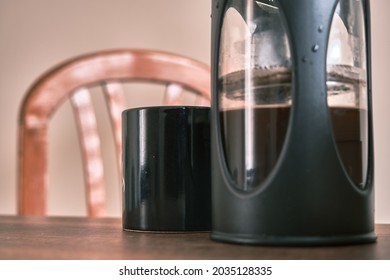 French Press Coffee Maker And It Is Half Filled With Arabic And Black Caffe Together And Also Chair With Black Caffe Pot With Upper Side Of Wooden Chair. All Things Standing On Wooden Table.