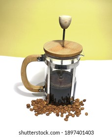 French Press Coffee Maker On The White Ground. Arabic Coffee Beans Bottom Of The French Press And Cooked Caffe Inside The Glass Side Of The Press And Wooded Details With Dull Yellow Background.