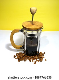 French Press Coffee Maker On The White Ground. Arabic Coffee Beans Bottom Of The French Press And Cooked Caffe Inside The Glass Side Of The Press And Wooded Details With Dull Yellow Background.