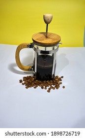 French Press Coffee Maker On The White Ground. Arabic Coffee Beans Bottom Of The French Press And Cooked Caffe Inside The Glass Side Of The Press And Wooded Details With Dull Yellow Background.