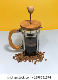 French Press Coffee Maker On The White Ground. Arabic Coffee Beans Bottom Of The French Press And Cooked Caffe Inside The Glass Side Of The Press And Wooded Details With Dull Yellow Background.