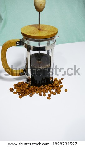 French press coffee maker on the white ground. Arabic coffee beans bottom of the french press and cooked caffe inside the glass side of the press and wooded details with dull green  background.