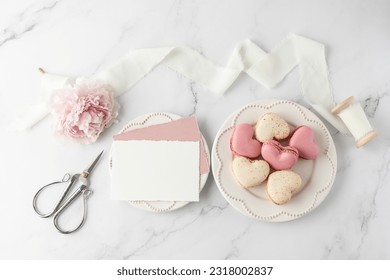 French sweet cookies macarons macaroons on retro plate and blank paper cards on marble table background. Heart shape macarons, peony, silk ribbon and decor. Mockup greeting card with copy space.