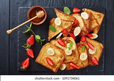 French toasts with appetizing, lacy brown crust and fluffy inside served on a black slate tray with strawberries, banana slices, mint and honey, view from above - Powered by Shutterstock