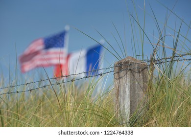 French and United States flags flying symbolically together over barbed wire against a blue sky over the sand dunes at Utah beach in Normandy  France signifying cooperation between France and the USA