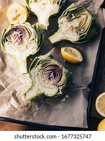 Fresh artichokes cut in halves on baking tray with lemons. Baking brown parchment paper. Baked artichokes. Healthy lunch time. Seasoning with salt. Direct sunlight. Purple and green artichoke hearts.