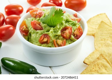 Fresh Avocado Guacamole Sauce Or Dip With Onion And Tomato In A White Bowl Over A Wooden Table With Corn Nachos Or Totopos, Tomatoes And Green Chili On The Side. Typical Mexican Appetizer Or Snack.