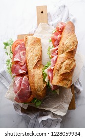 Fresh baguette sandwich bahn-mi styled. Ham, sliced cheese, tomatoes and fresh lettuce on wooden cutting board on white marble background.