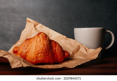 Fresh Baked Croissant On Craft Paper, Mug Of Coffee On Wooden Table. Cup Of Hot Drink, Bun In Wrap Close Up. Food, French Breakfast, Bakery, Morning Menu, Cafe Concept. Side View, Copy Space