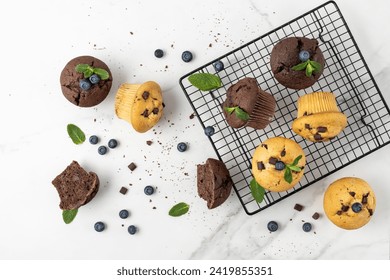 Fresh baked muffins with chocolate chips, blueberry berries and mint leaves on baking rack and on white marble table background. Chocolate  cupcake closeup as dessert for coffee break. Top view.