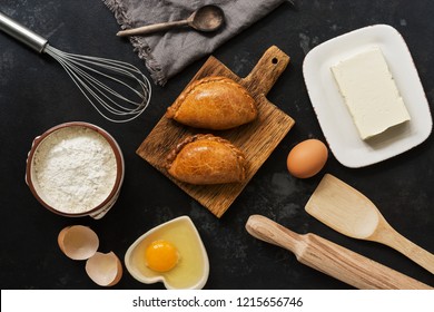 Fresh baked pasties and baking ingredients on a dark stone background. Russian pirozhki. Top view. - Shutterstock ID 1215656746