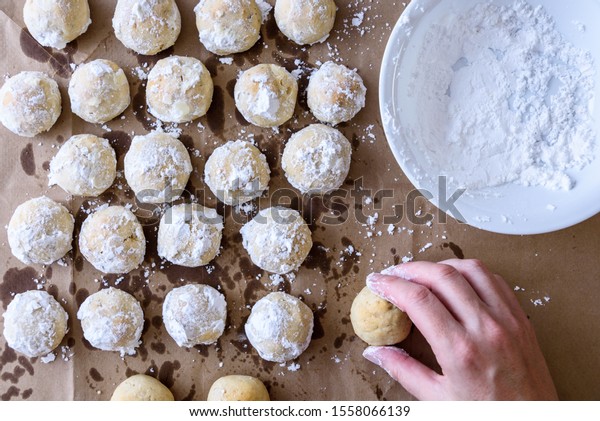 Fresh baked Russian Tea Cake cookies cooking on\
brown paper, woman’s hand, bowl of powdered sugar ready to roll\
cookies in\
