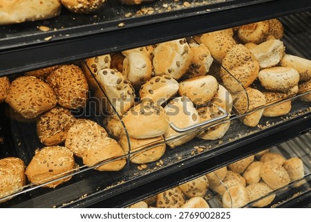 Fresh bakery rolls and seeded bread displayed on store shelves behind glass. Assorted buns in supermarket bakery section show daily baked food products for sale.