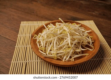 Fresh bean sprouts fill a rustic wooden bowl. They rest on a woven bamboo mat beside a dark wood table. Delicate white stems and green tips show natural texture. The scene evokes freshness, health
