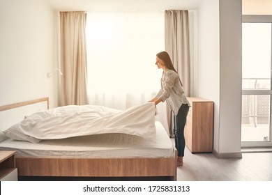 Fresh Bedclothes. Pretty Female Person Keeping Smile On Her Face While Making Her Bed