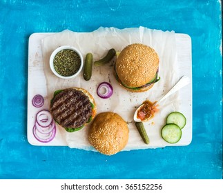 Fresh Beef Burger With Cheese, Vegetables And Spicy Tomato Sauce On Paper And White Serving Board, Blue Wooden Background, Top View