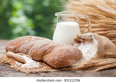 Fresh bread, jug of milk, sack with flour and sheaf of wheat on wooden table.