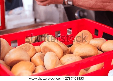 Fresh bread rolls are stacked in a red plastic basket at a food market, with a vendor's hand preparing items in the background