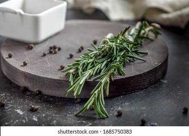 Fresh bunch and flower of rosemary and pepper in a white bowl on a wooden board on a background of dark concrete table and a beige towel. Herbs and spices.
