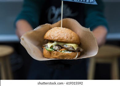 Fresh burger cooked at barbecue outdoors in craft paper. Big hamburger with steak meat and vegetables closeup, chef unfocused at background. Street fast food.