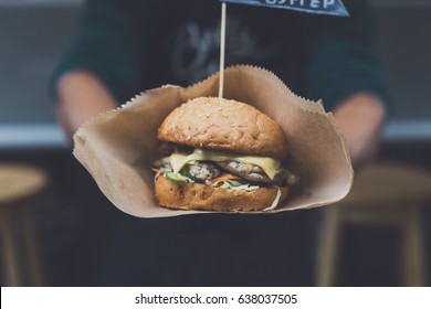 Fresh Burger Cooked At Barbecue Outdoors In Craft Paper. Cookout American Bbq Food. Big Hamburger With Steak Meat And Vegetables Closeup With Chef Unfocused At Background. Street Food, Fast Food.