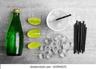 Fresh cocktail preparation: soda bottle, ice cubes, slices of lime  on grey table background, top view