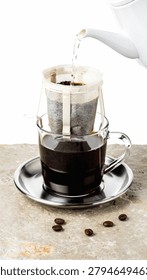Fresh coffee being brewed using a drip bag coffee filter attached to a glass mug while hot water is poured from a white kettle.