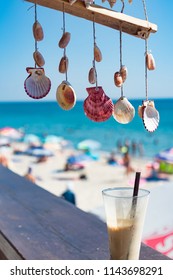 Fresh cold coffee ice milk frappe drink in transparent glass with colorful sea shells hamging on twine string as summer decoration. Blurred background of blue sky, sea water, sunny beach. Relaxation.