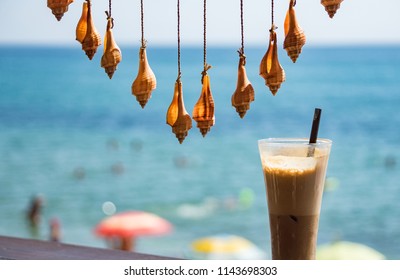Fresh cold coffee ice milk frappe drink in transparent glass with colorful sea shells hamging on twine string as summer decoration. Blurred background of blue sky, sea water, sunny beach. Relaxation.