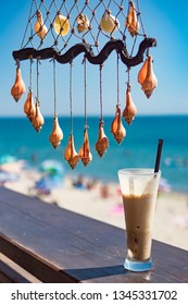 Fresh cold coffee ice milk frappe drink in transparent glass with colorful sea shells hanging on twine string as summer decoration. Blurred background of blue sky, sea water, sunny beach. Relaxation.
