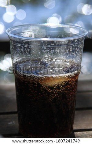A fresh and cold cola drink with three ice cubes in plastic mug on a blurry, dark brown wooden table, sparkling all the way in the mug, 