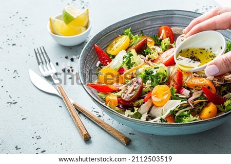Fresh colorful spring vegetable salad with cherry tomatoes and sweet peppers in the blue bowl. Cook’s hand pouring olive oil with herbs. Healthy organic vegan lunch or appetizer close up.