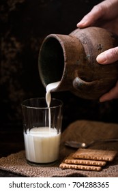Fresh Cow Milk Poured Into Glass From Clay Jug In Female Hands, Butter Cookies Stack On Sack Cloth And Spoon By Side, Rural Scene In Rustic Style And Dark Background, Organic Breakfast, Copy Space
