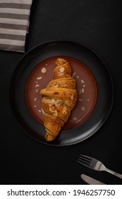 Fresh Croissant With Almond Butter In Dark Plate And Cup Of Coffee In Colored Napkin  On Black Background
