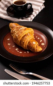 Fresh Croissant With Almond Butter In Dark Plate And Cup Of Coffee In Colored Napkin  On Black Background
