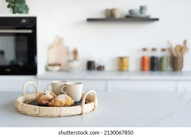 Fresh croissants and cups of tea in a basket in contemporary kitchen interior. Kitchen appliances and decor on background. Homemade bakery concept. Modern white furniture