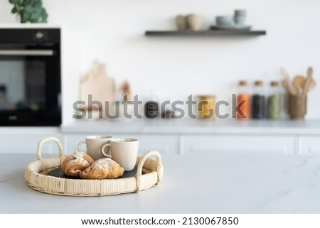 Fresh croissants and cups of tea in a basket in contemporary kitchen interior. Kitchen appliances and decor on background. Homemade bakery concept. Modern white furniture