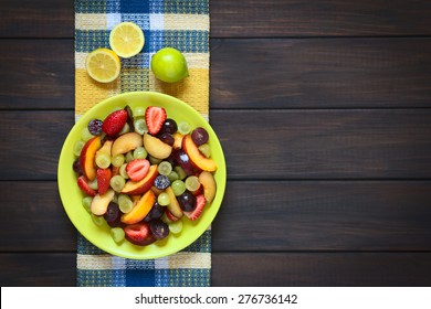 Fresh Fruit Salad Made Of Grape, Strawberry, Plum And Nectarine Served On Plate With Lemon Above, Photographed Overhead On Dark Wood With Natural Light