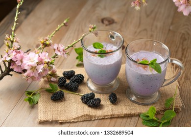 Fresh Fruit Smoothie Blackberry Milkshake, Purple Yogurt Or Ice Cream Drink On Wood Table With Sprigs Of Mint, Blackberries, And Cherry Blossoms. Springtime Healthy Shake Recipe In Beautiful Setting.