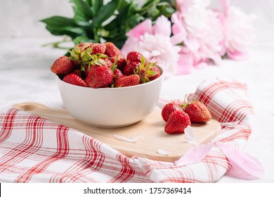 Fresh garden strawberry in a plate on a wooden kitchen board with a beautiful napkin and serving, wholesome food and vitamins, diet. Pink and white peony flowers, side view