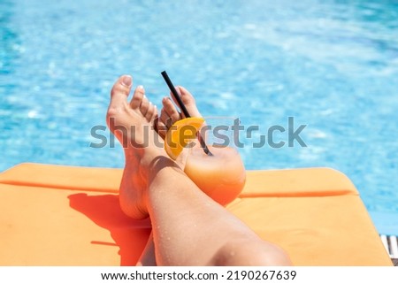 Fresh glass of a cocktail with an orange slice and woman legs on a sun lounger by a swimming pool. Holiday and vacation concept.
Selective focus.