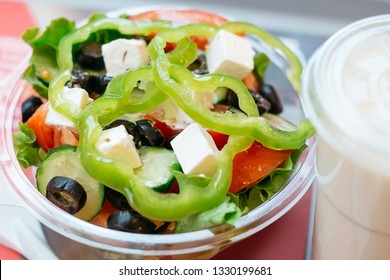 Fresh Greek Salad: Pepper, Tomato, Olive, Feta Cheese, Cucumber, Lettuce, Oil.   To Go Salad In A Transparent Bowl Near A Drink On A Table. Close Up, Detailed Image.