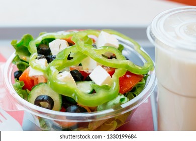 Fresh Greek Salad With Pepper, Tomato, Olive, Feta Cheese, Cucumber, Lettuce, Oil. To Go Salad In A Transparent Bowl Near A Drink On A Table. Close Up.