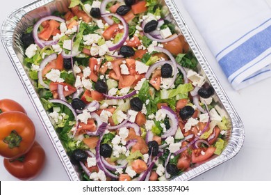 A Fresh Greek Salad With Romain Lettuce, Tomatoes, Olives, Feta Cheese And Red Onion Slices In A Catering Style Aluminium Tray And Next To It A Napkin With A Blue Stipe And Some Whole Fresh Tomatoes.